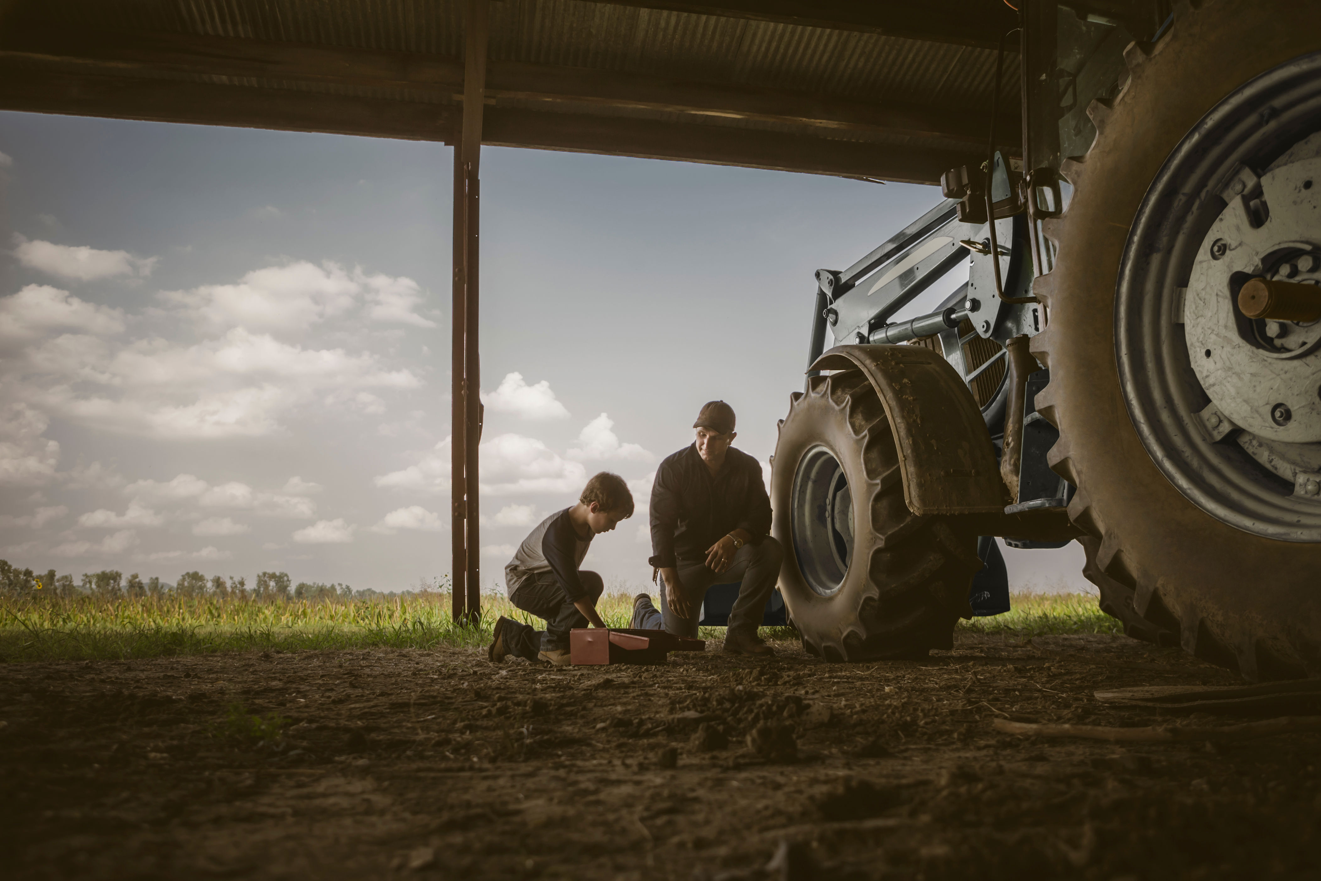 A child and an adult kneel next to a tractor in a barn, focusing on a toolbox, with a field visible in the background