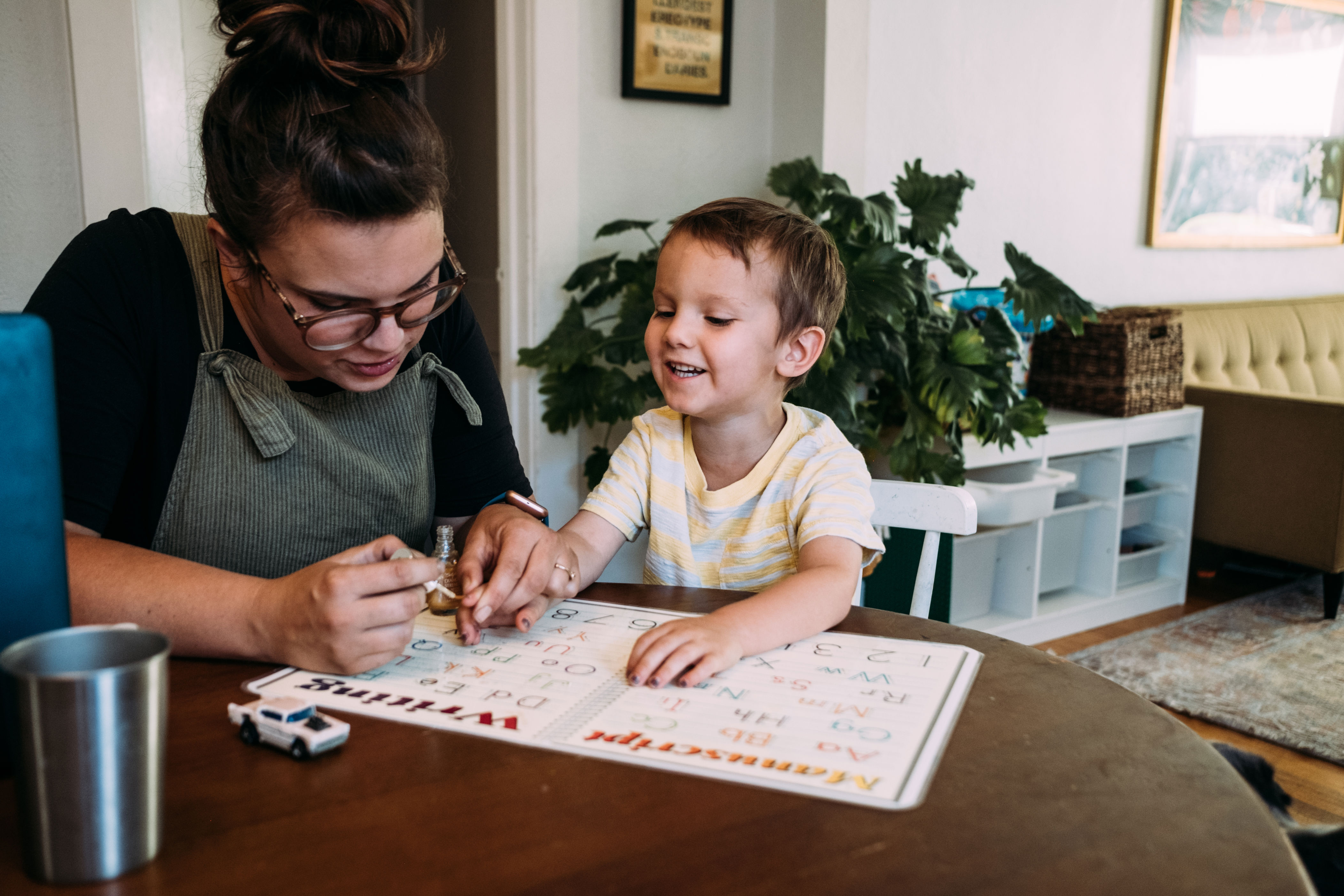 Adult helping a child with a learning activity at a table in a cozy living room. A toy car is on the table