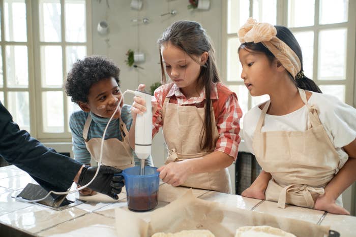Three children wearing aprons use a hand blender together in a kitchen, guided by an adult's hand
