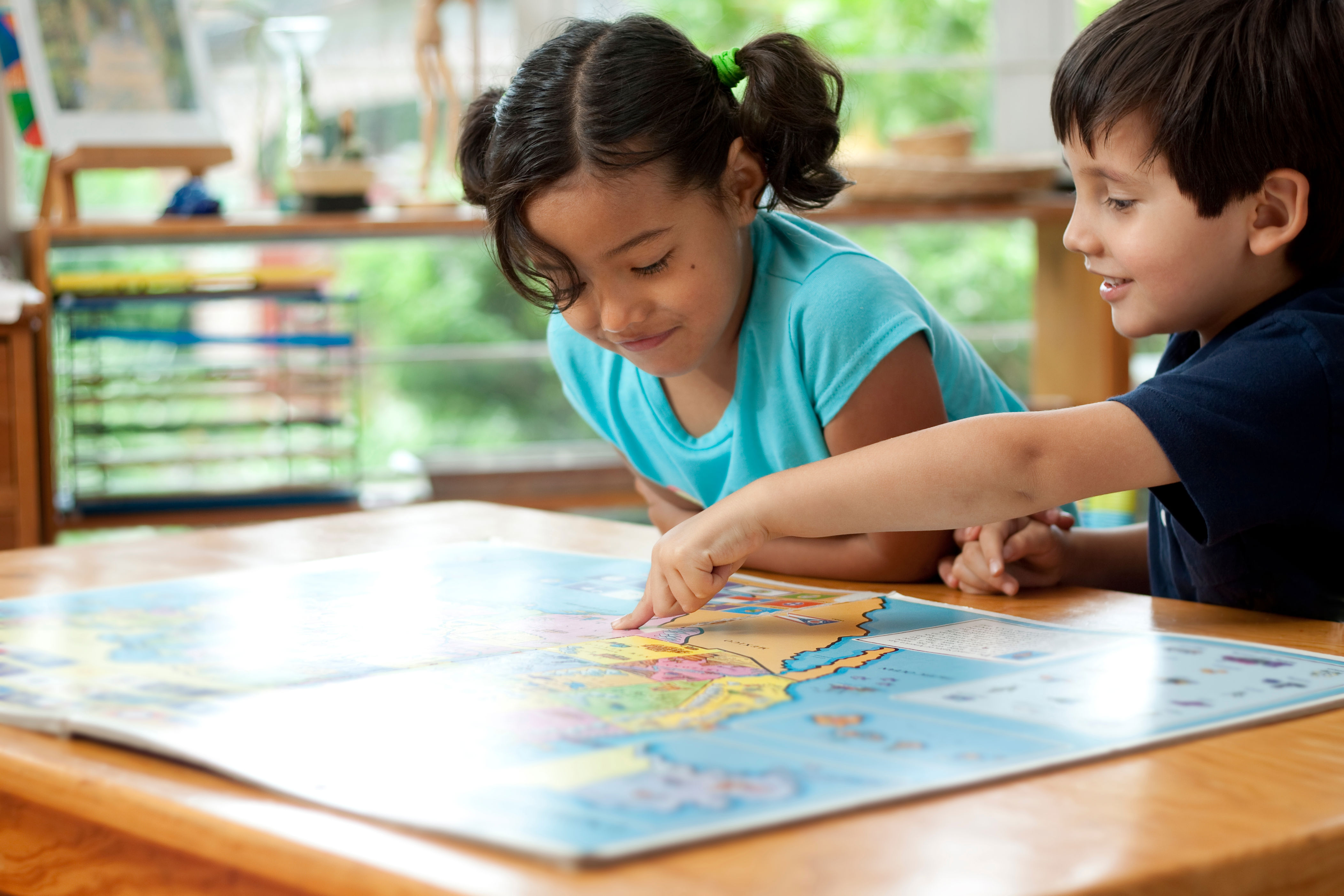 Two children happily explore a world map on a table, pointing and discussing different locations