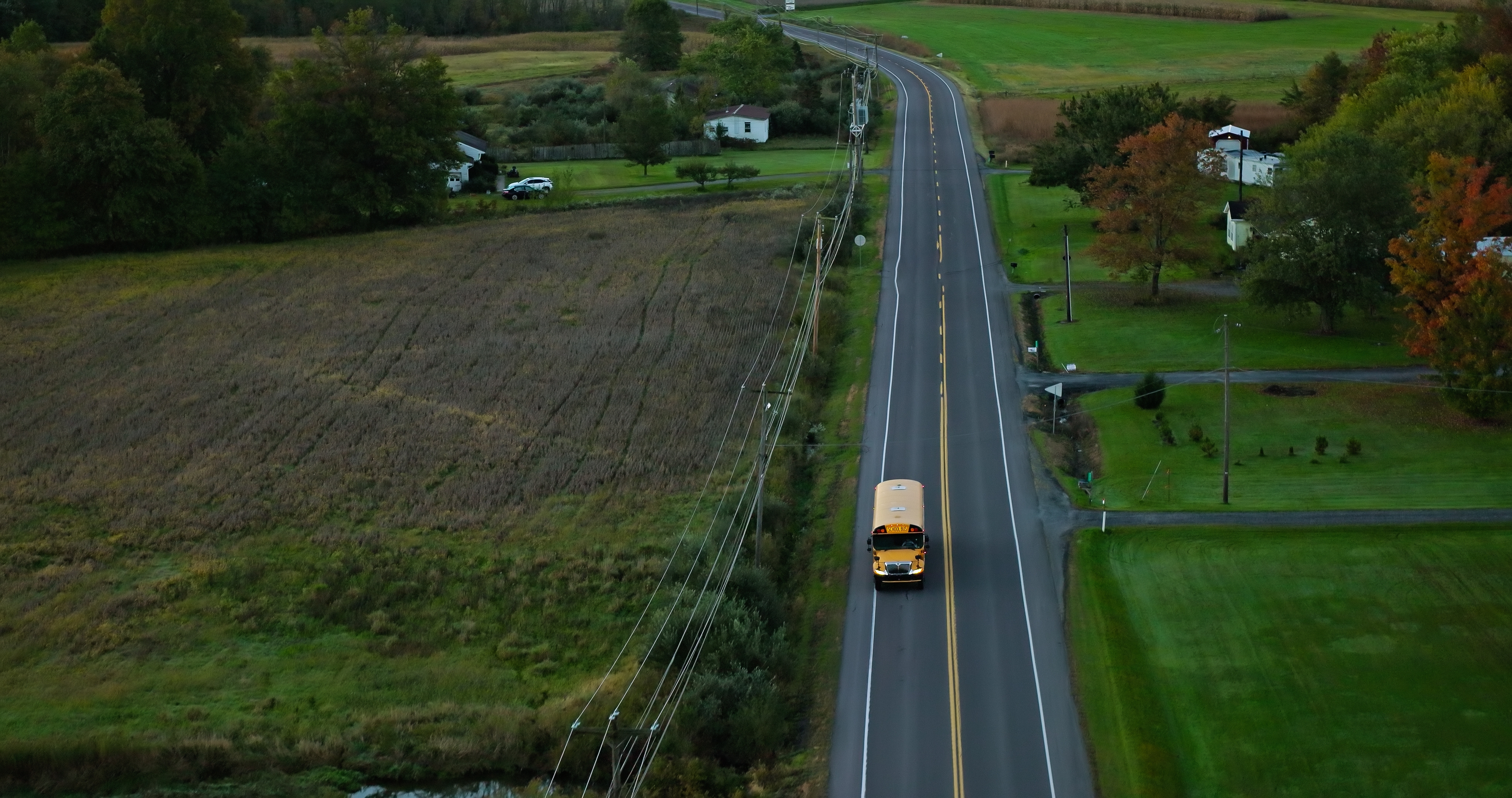 Aerial view of a rural road with a yellow school bus traveling through green fields and houses in the distance
