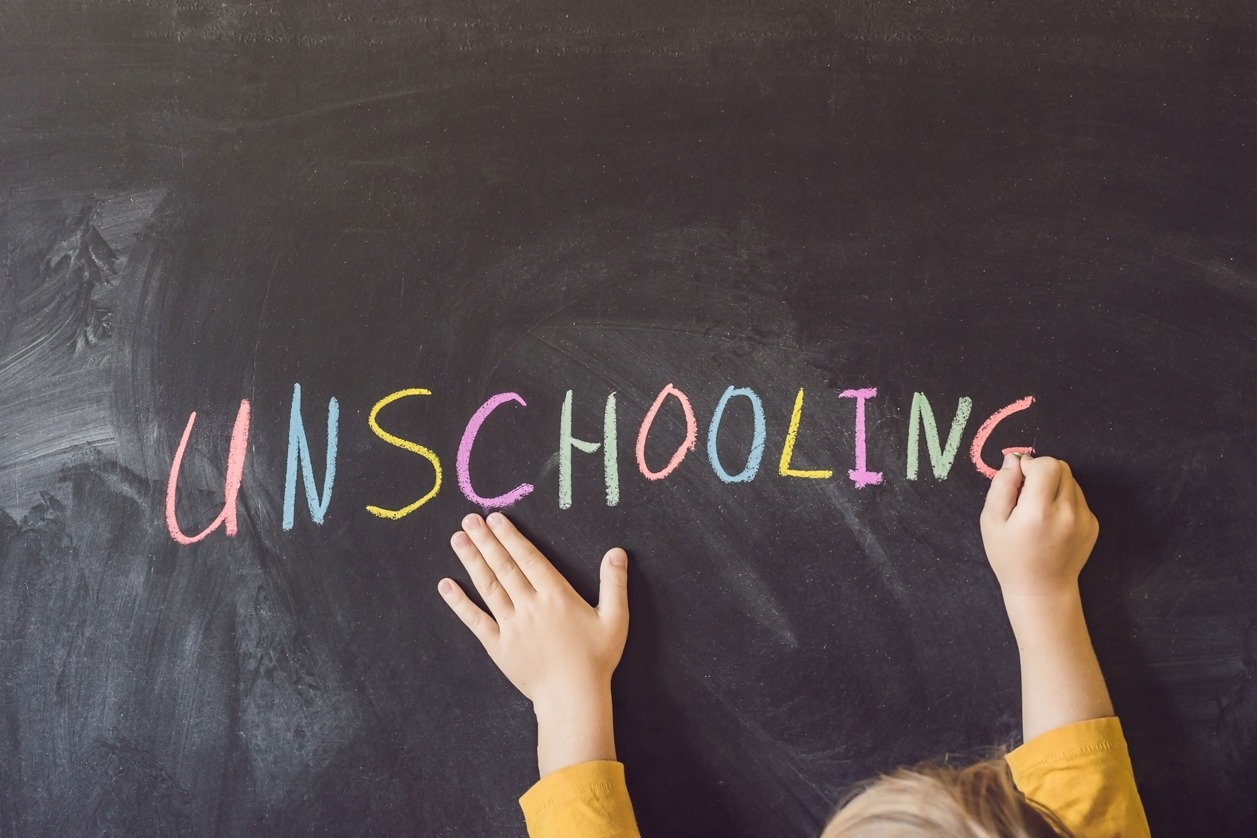Child writing "UNSCHOOLING" on a chalkboard with colorful chalk