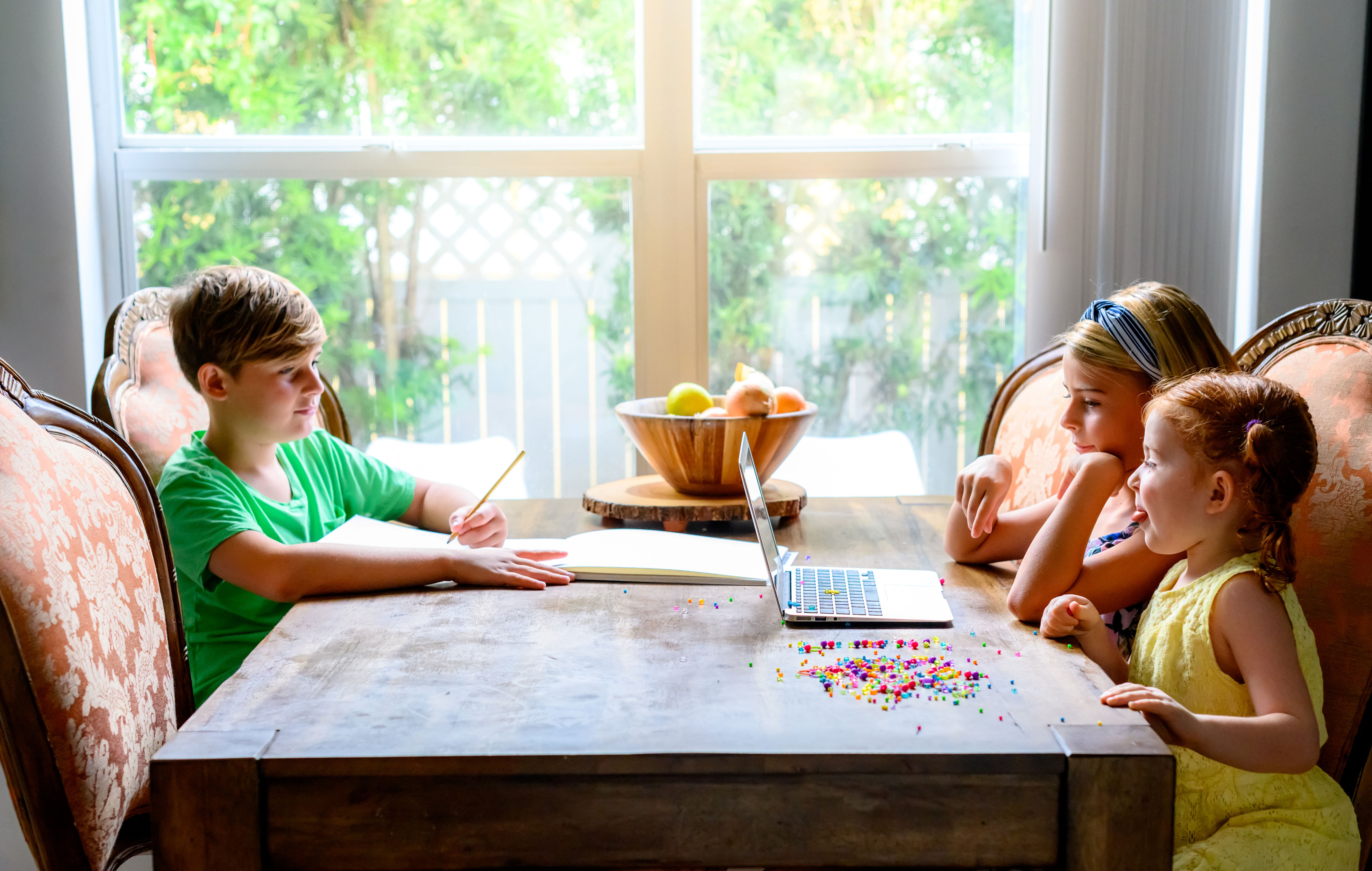 Three children sit at a table with a laptop and colorful beads, appearing to collaborate on a project