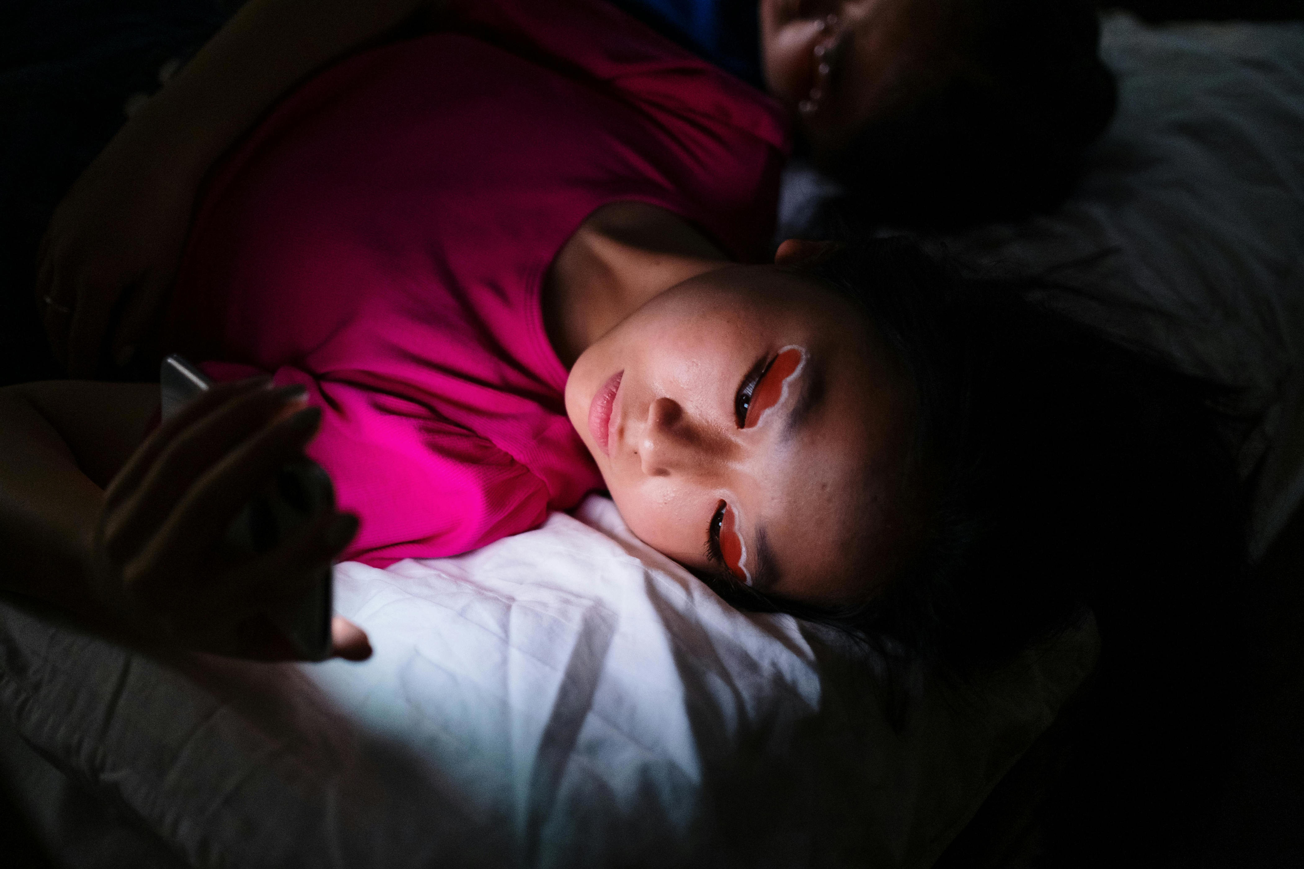Asian woman lying in bed using smartphone in dark room, illustrating digital distraction.