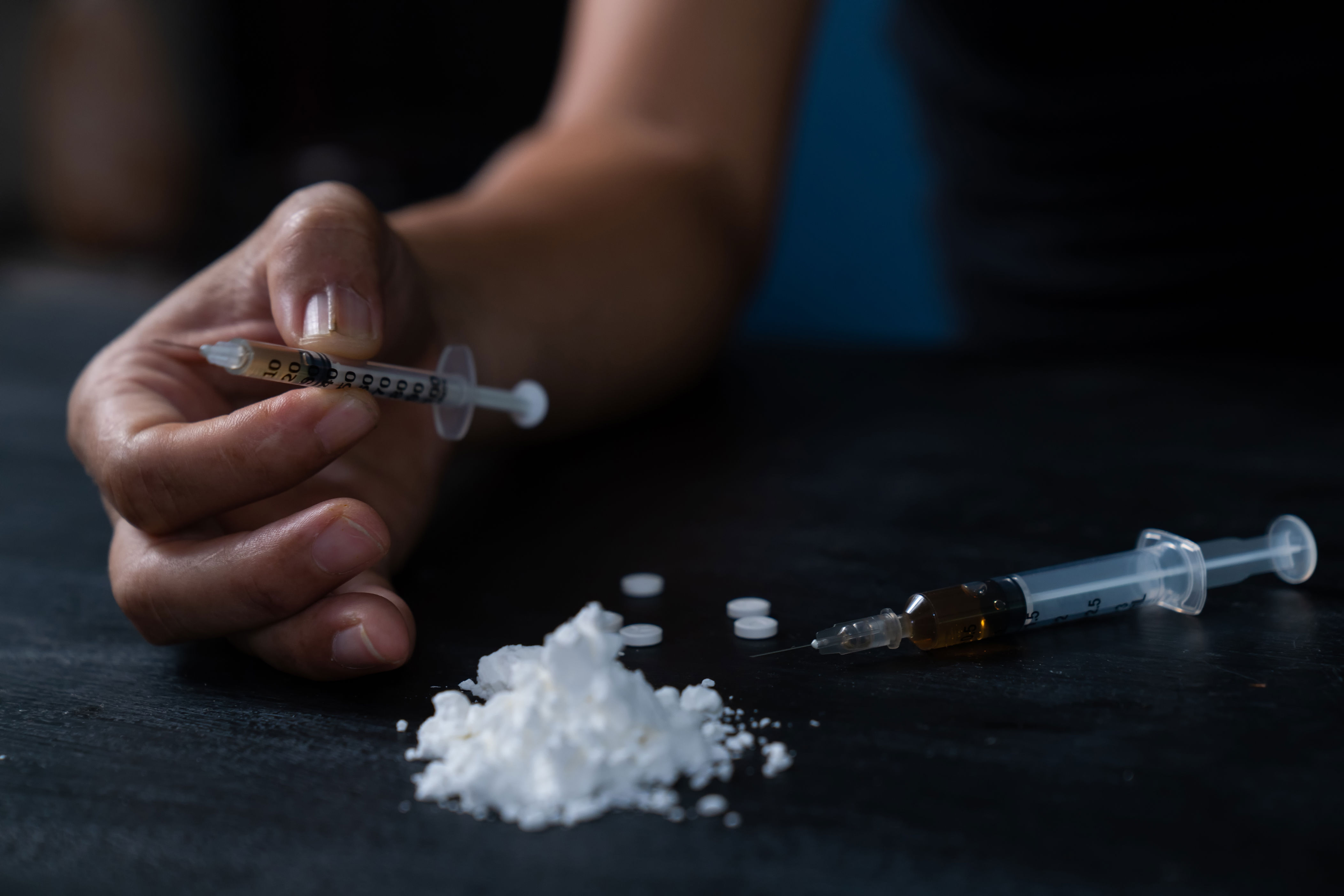 A hand holding a syringe, next to a syringe, powder, and pills on a dark surface