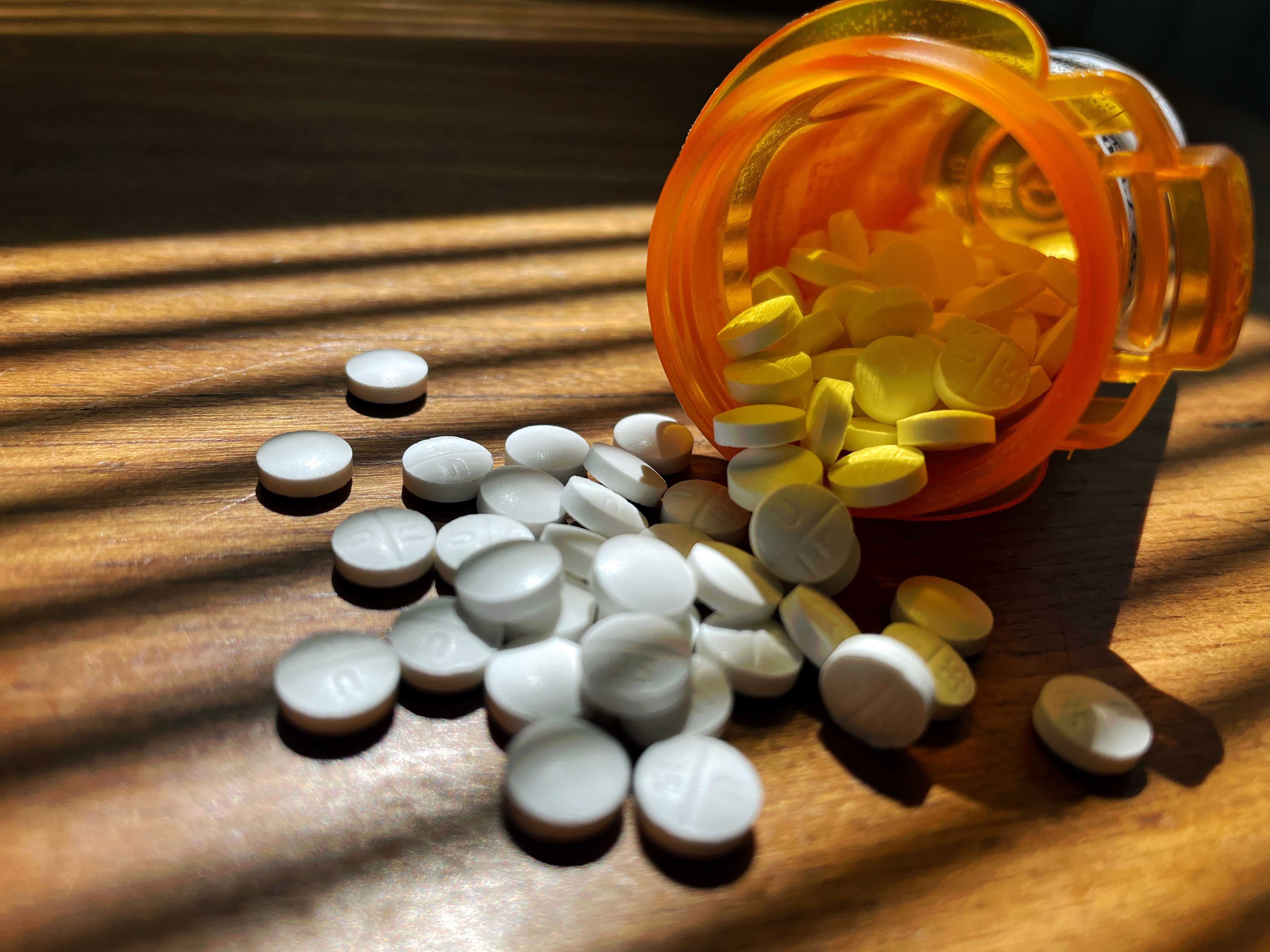 Pills spilling from an orange prescription bottle onto a wooden surface, with sunlight casting shadows across the scene