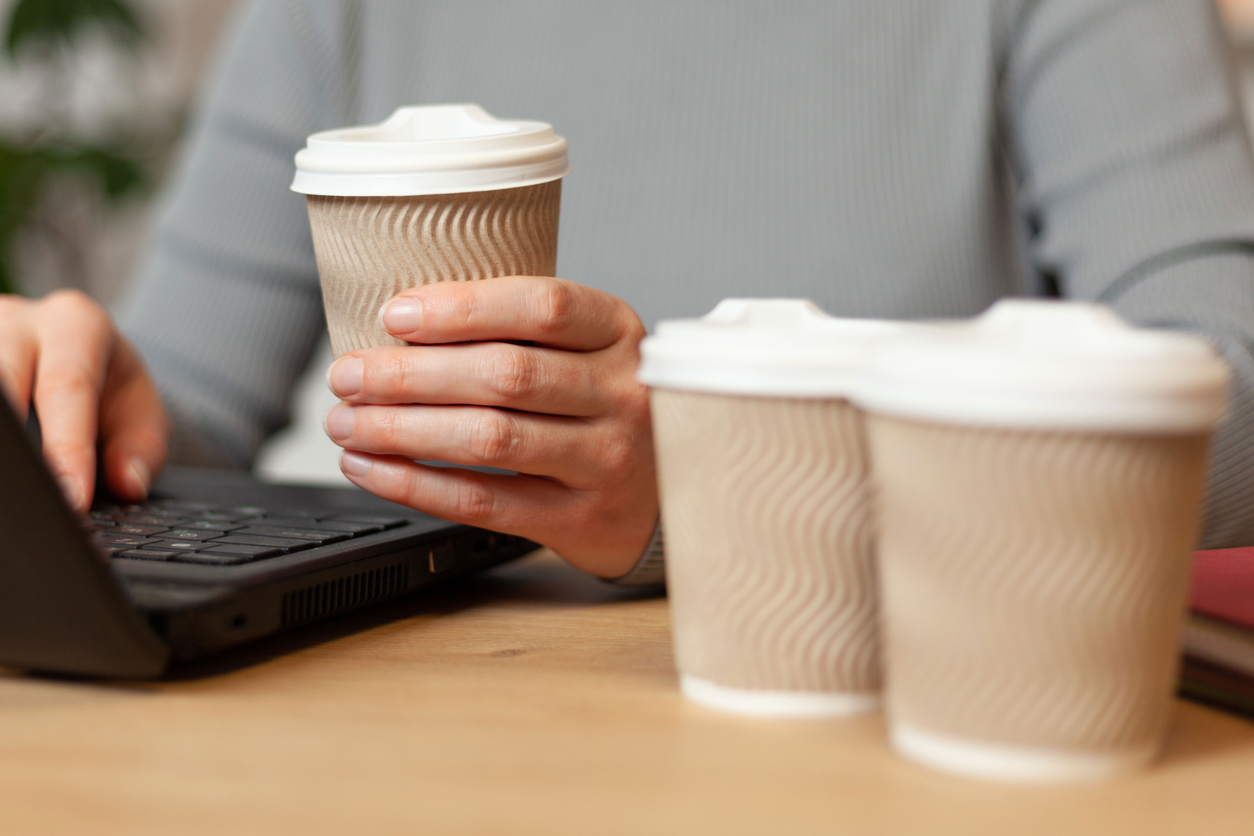 Person holds a to-go coffee cup while typing on a laptop, with two more cups nearby on a table