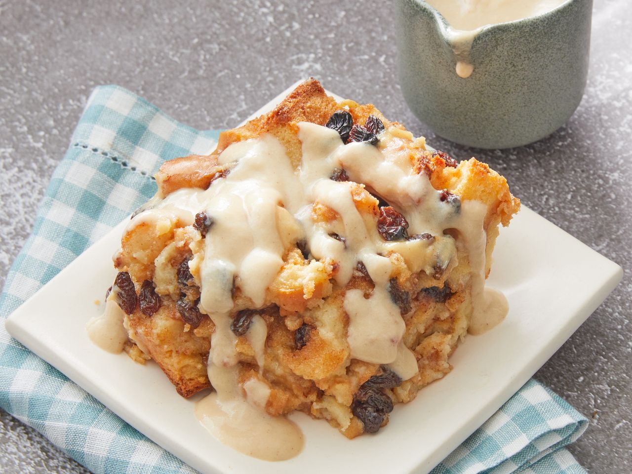 A slice of bread pudding with vanilla sauce served on a plate, a creamy pitcher in the background