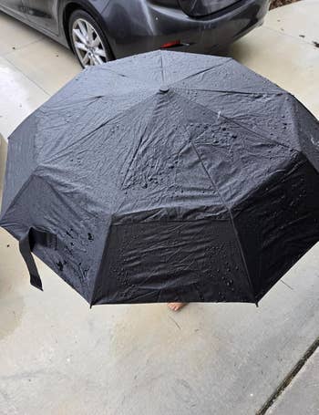 A black umbrella with rain droplets on it is open on a sidewalk near a parked car
