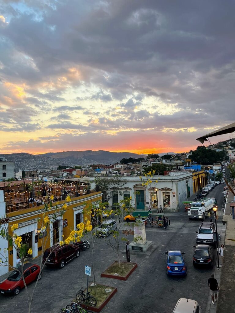Colorful buildings line a cobblestone street in Oaxaca at sunset, with yellow trees and a rooftop restaurant glowing under string lights as the sun sets behind the hills.