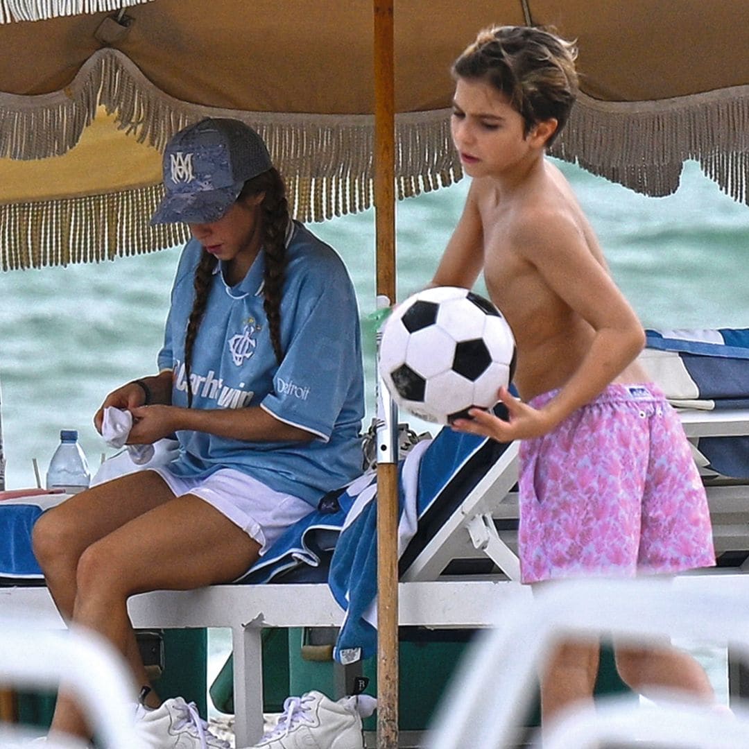 Shakira en la playa sentada bajo la sombrilla mientras su hijo pequeño juega con pelota de fútbol en la arena 