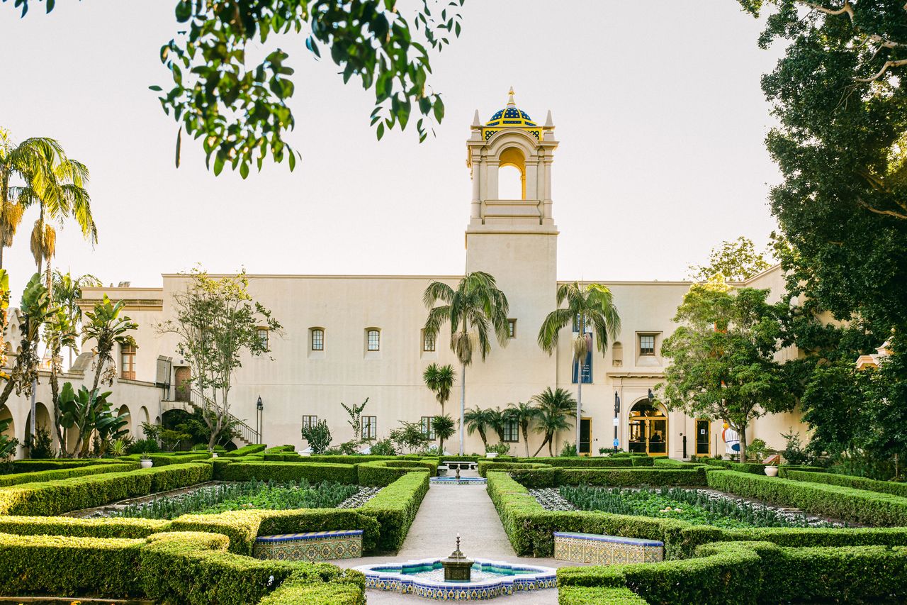 A manicured garden with a walking path leads to a building with a tall bell tower surrounded by palm trees