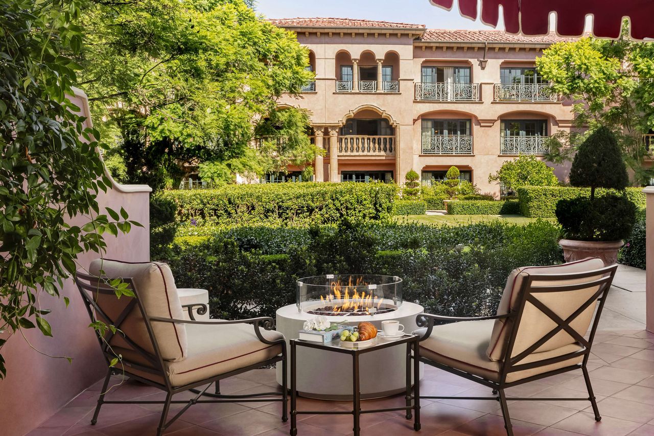 Two chairs with cushions surrounding a small fire feature in a courtyard with a luxury hotel facade and manicured gardens in the background