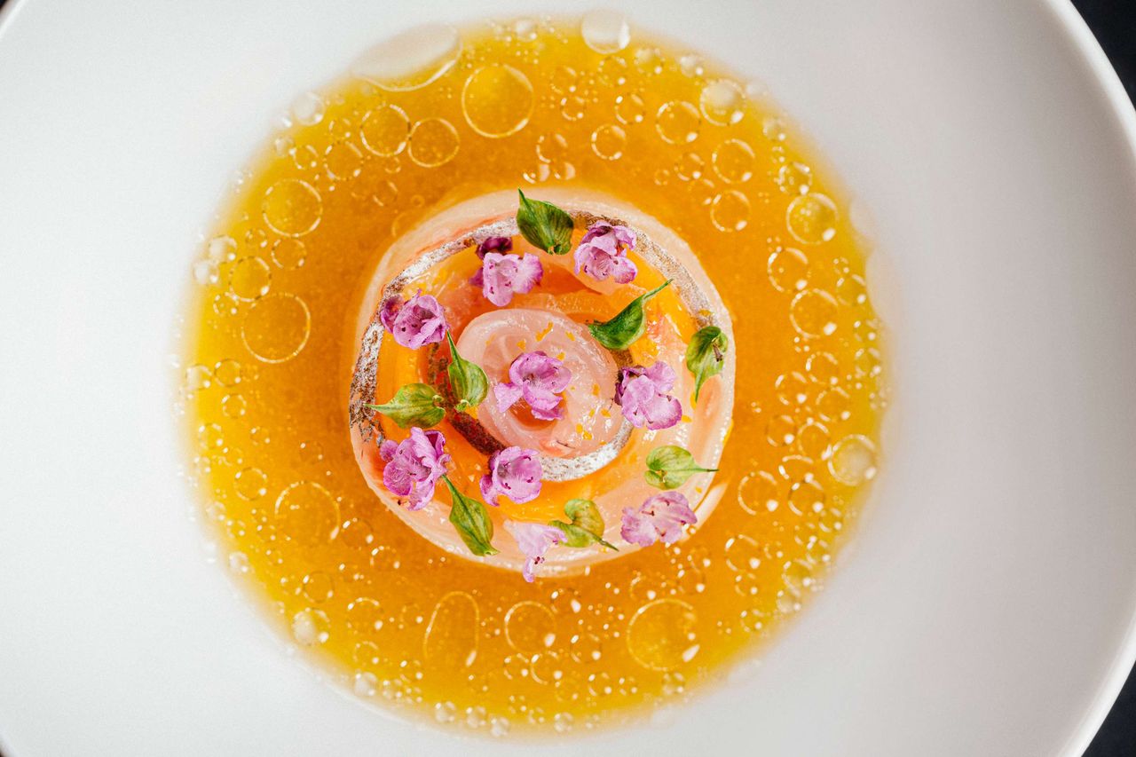 A gourmet dish with a circular arrangement of food garnished with small flowers and herbs served on a yellow sauce base