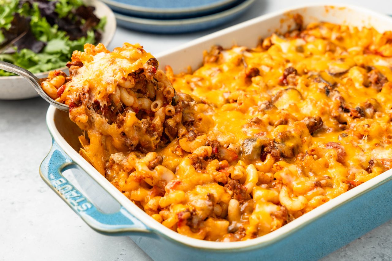 Spoonful of Johnny Marzetti Casserole lifted from the casserole dish, and in the surroundings, a stack of plates and a bowl of mixed greens salad