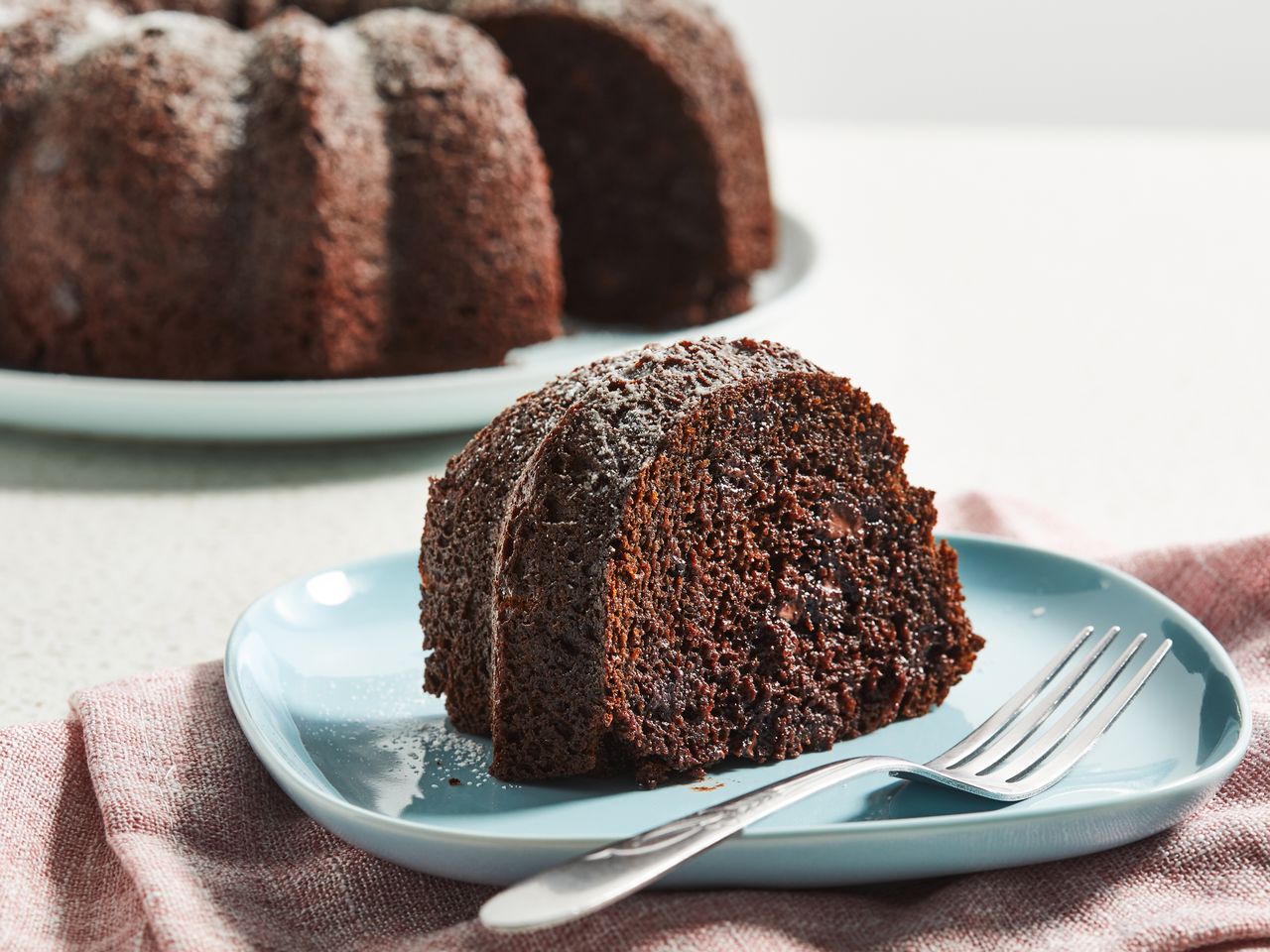 close up view of a slice of chocolate cake on a blue plate with a fork, and chocolate cake on a platter in the background