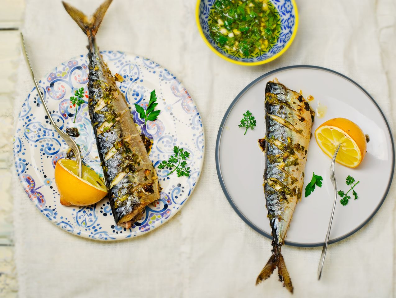 Two plates of grilled fish garnished with lemon and herbs accompanied by a small bowl of sauce on a white surface