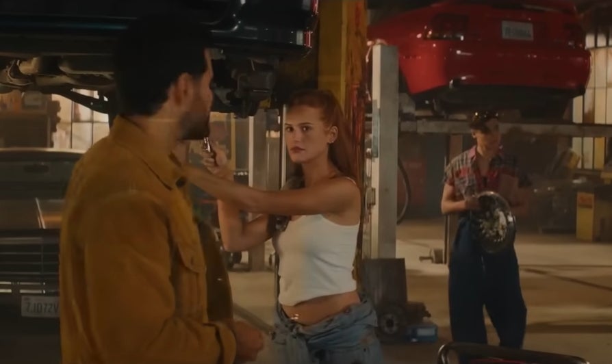 Two people in a garage, engaged in conversation. One wears casual workwear; the other, a sleeveless top and jeans. Another person works on a car in the background