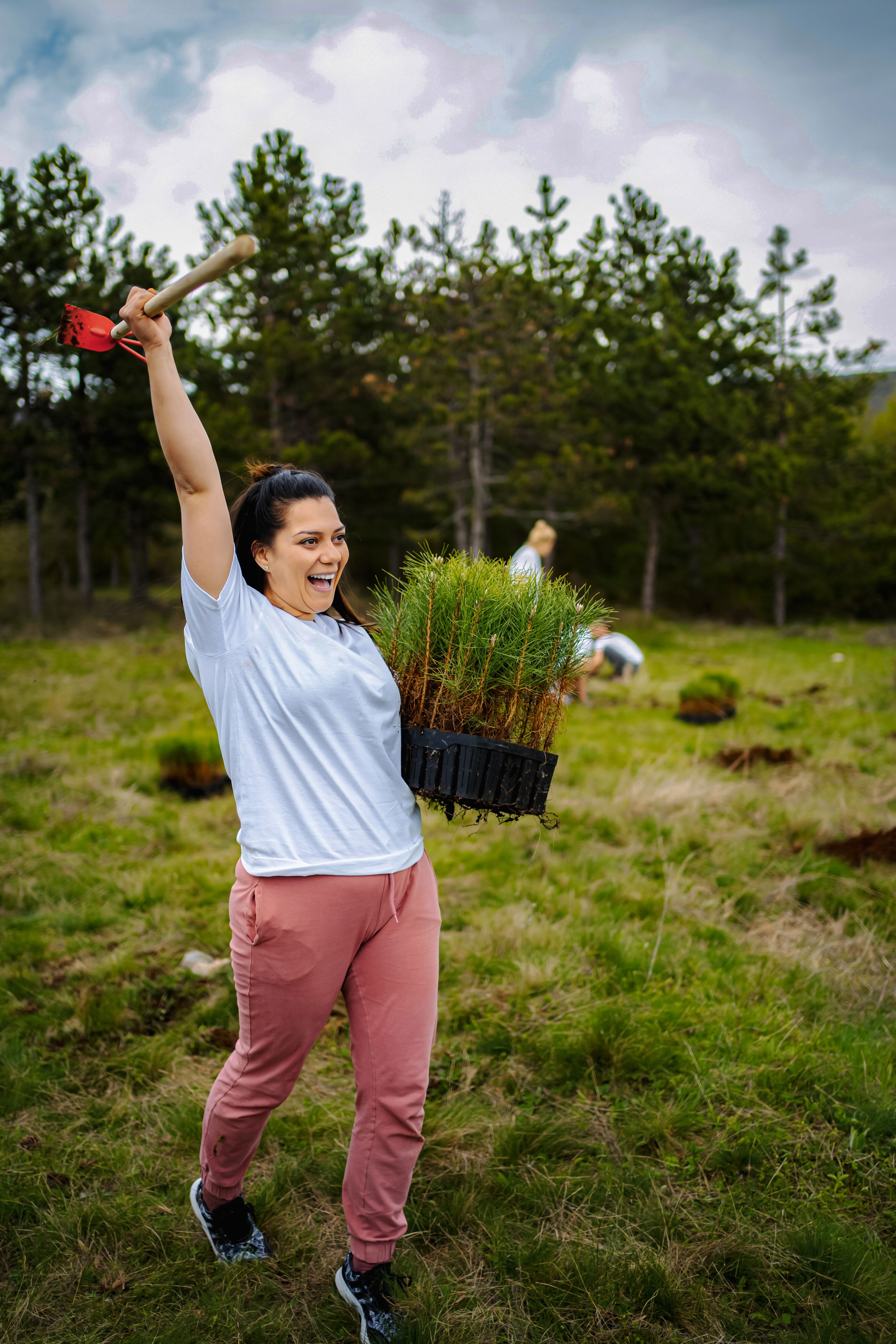 A person joyfully carries a tray of small plants and a shovel, participating in a tree-planting event in a grassy field with pine trees in the background