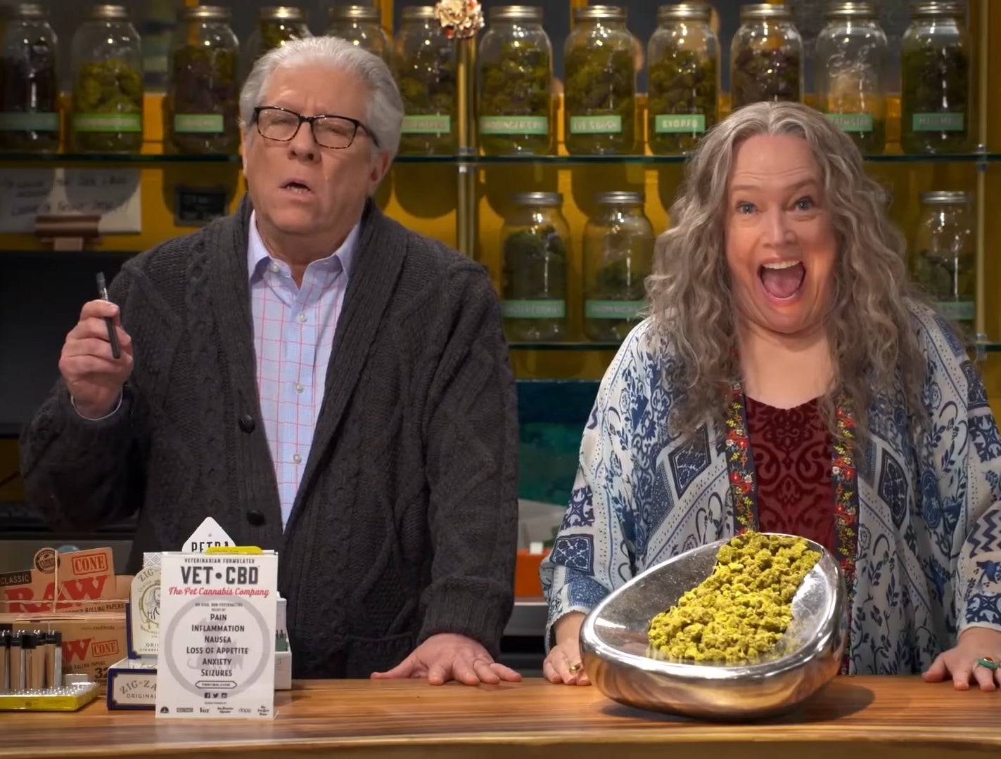 Older man and Kathy Bates in in a weed shop with jars behind them in "Disjointed" One looks surprised, the other thoughtful. A large bowl of hemp is on the counter.