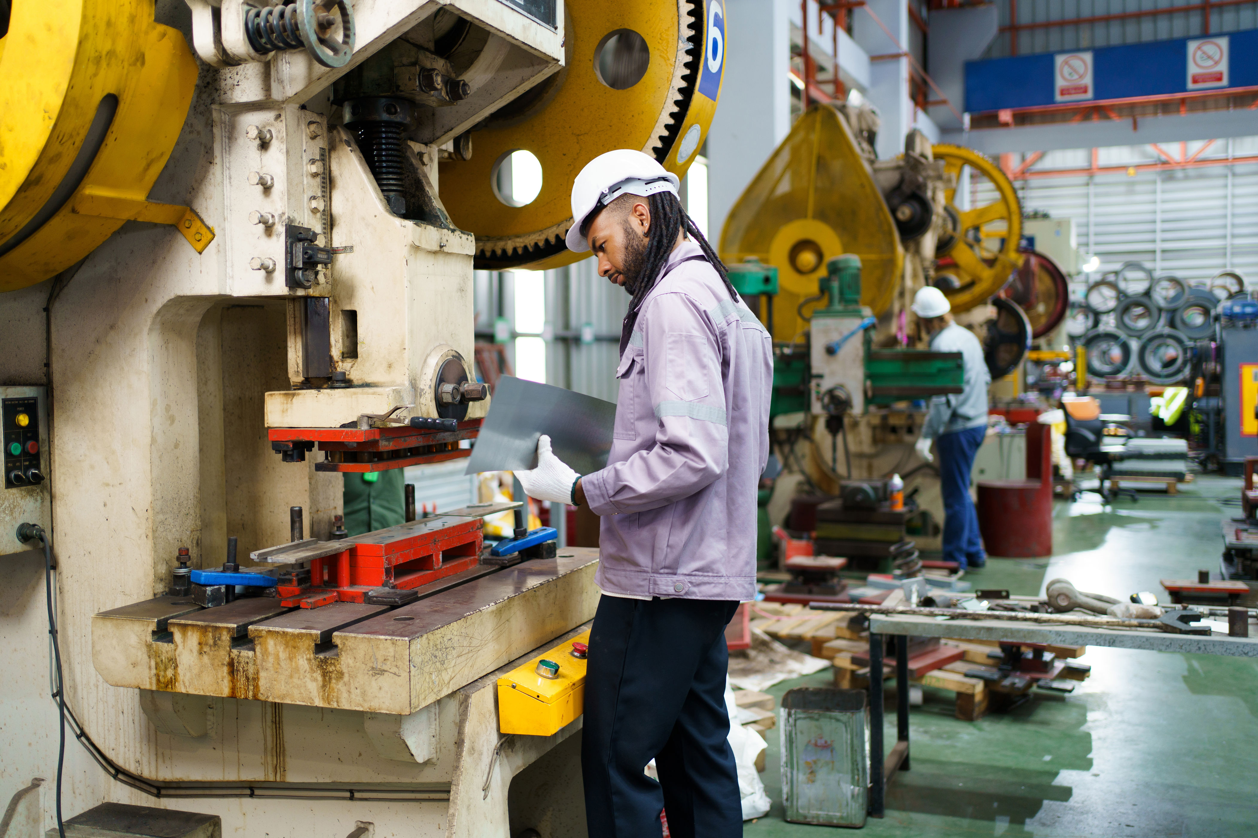 A worker wearing a hard hat and safety gear operates an industrial machine in a busy factory setting