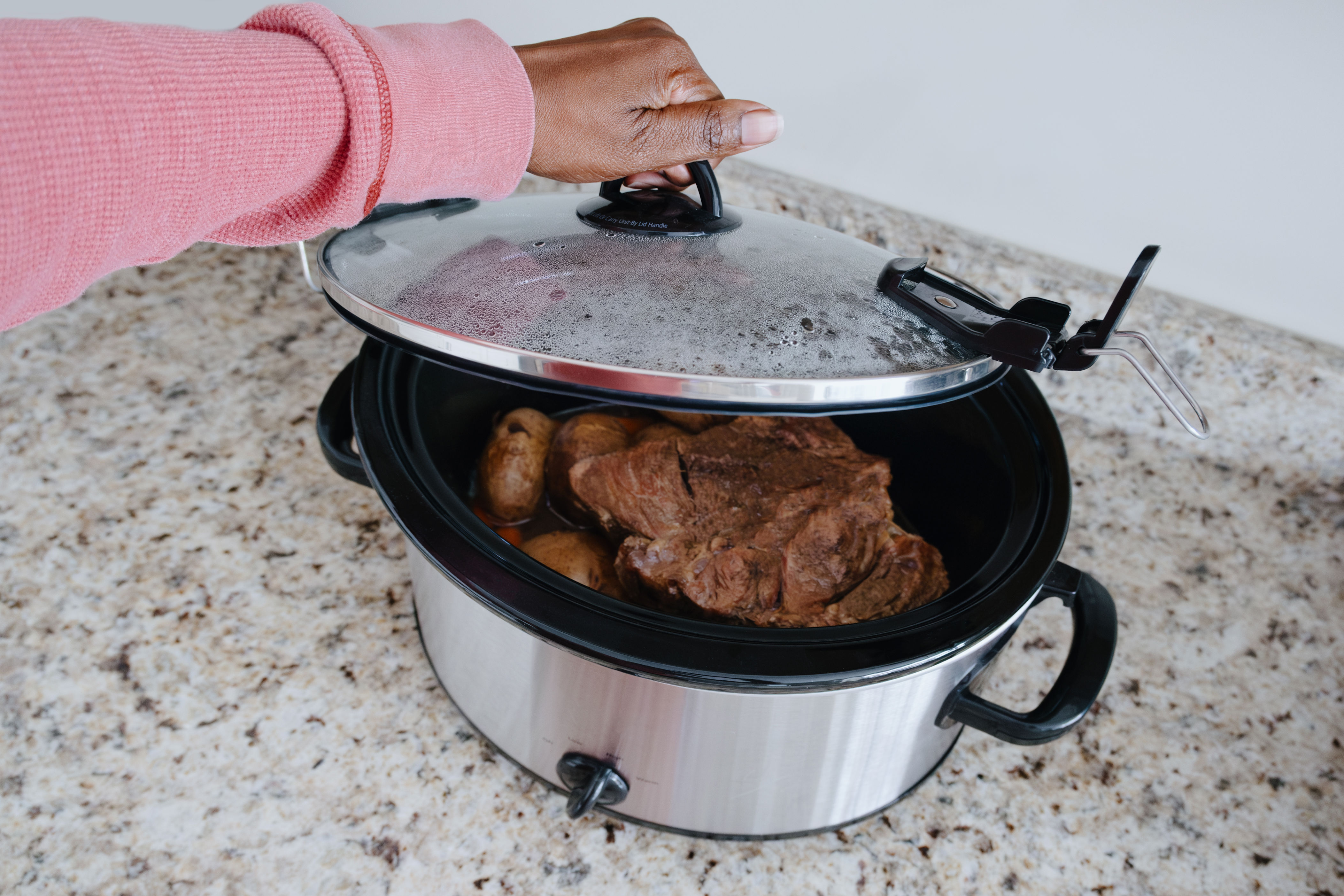 Hand lifting lid of a slow cooker containing a cooked roast and potatoes on a kitchen counter