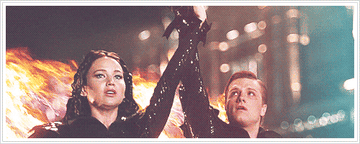 Jennifer Lawrence and Josh Hutcherson raise clasped hands triumphantly, wearing sleek outfits, set against a dramatic backdrop of flames in "The Hunger Games"