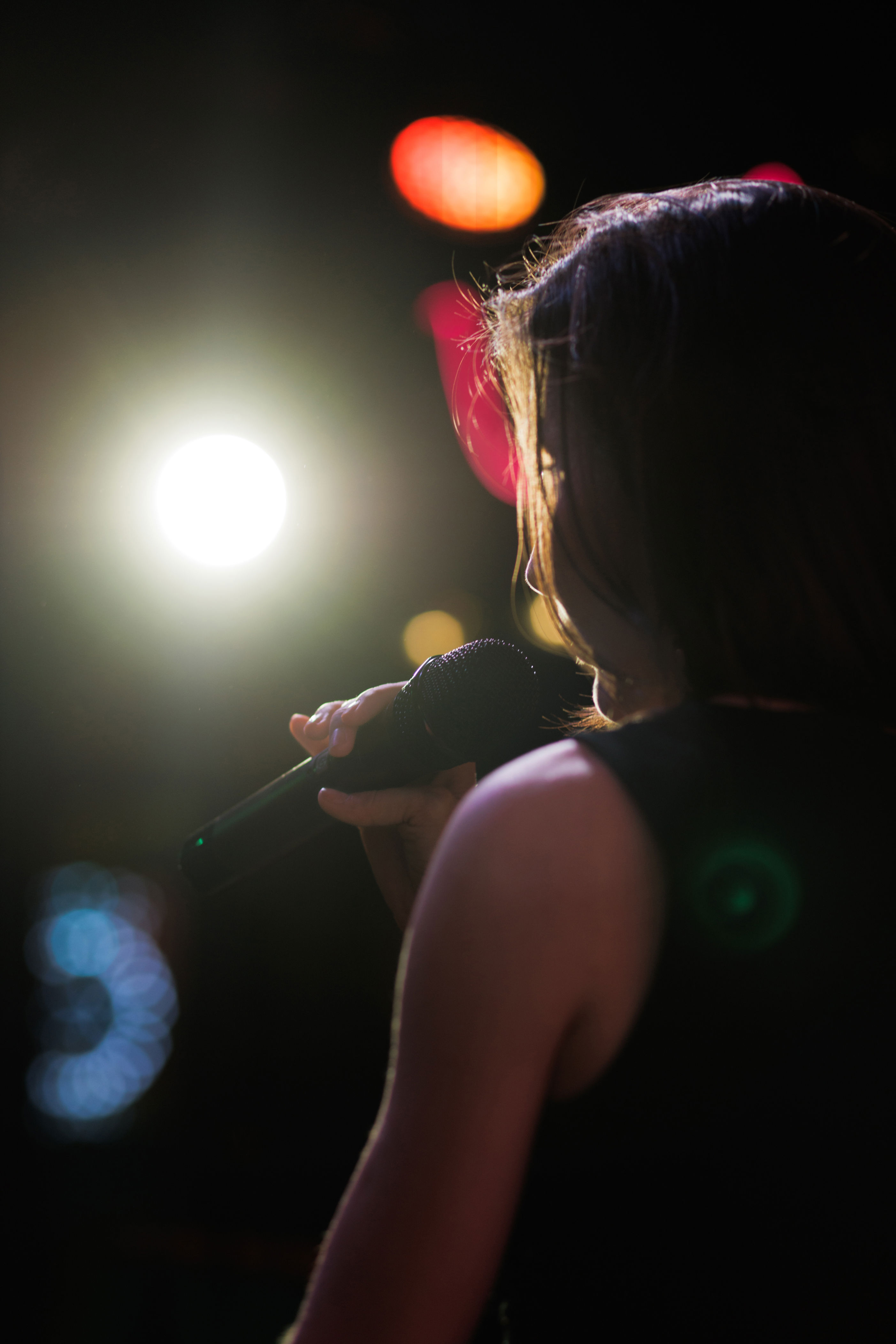 Person holding a microphone on stage with bright lights, captured from behind