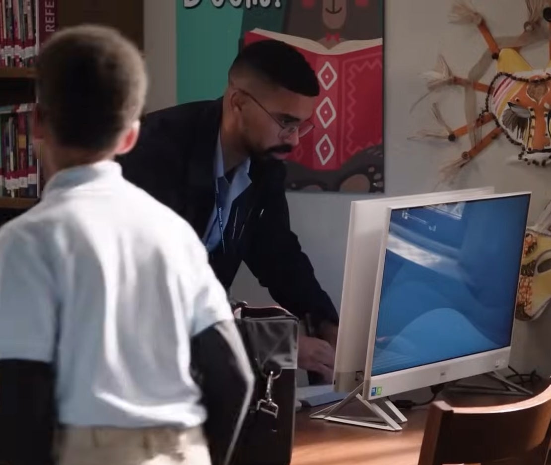 A teacher assists students in a library with a computer on a wooden desk. A wall decoration is in the background in "Abbott Elementary"
