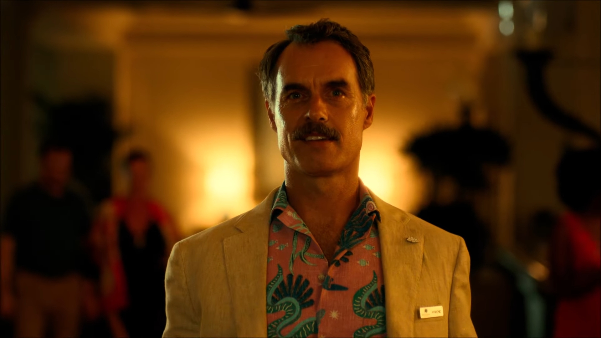 Person in a patterned shirt and light blazer at an event, wearing a name badge, possibly signifying a professional setting in "The White Lotus"