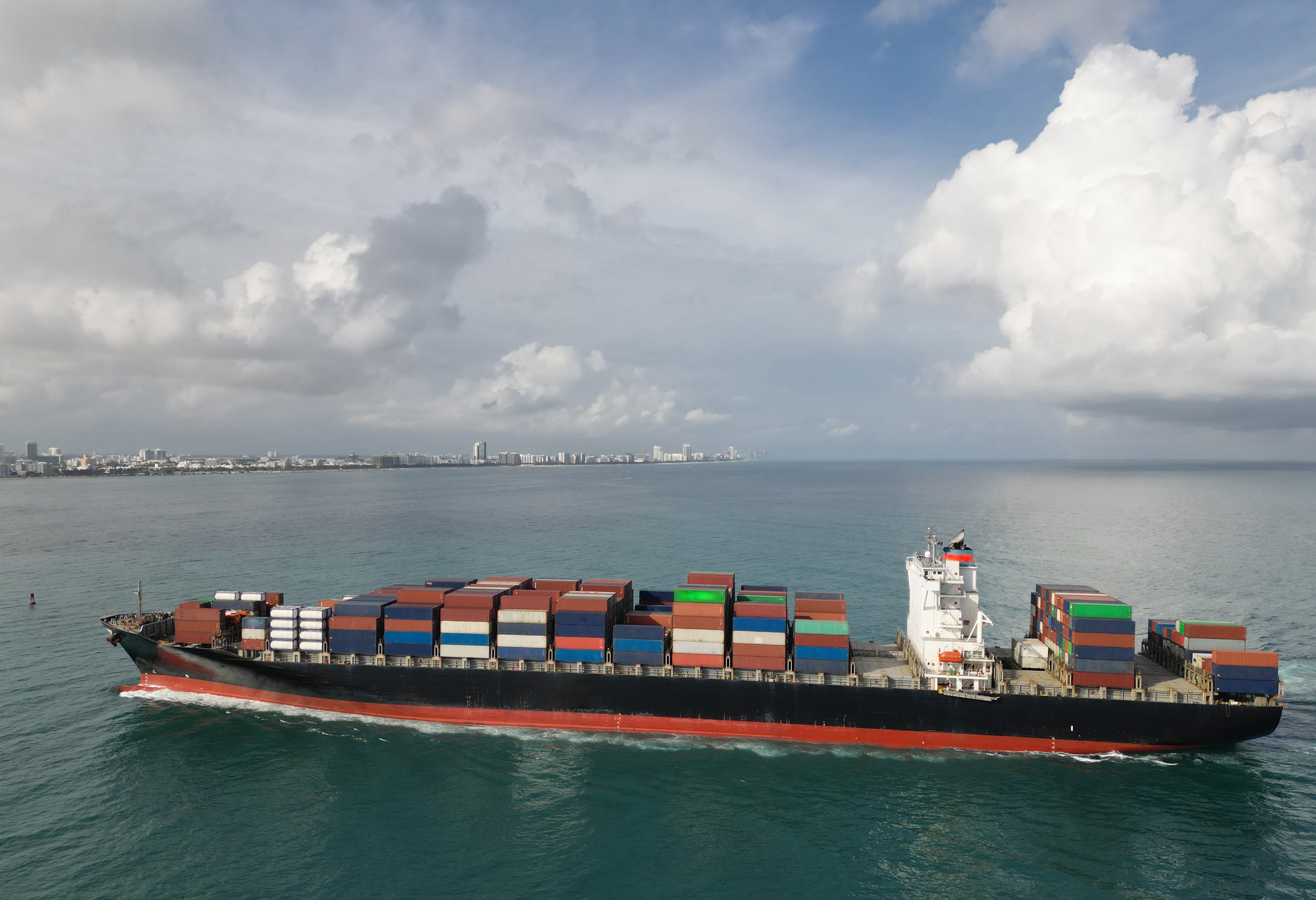 Large cargo ship transporting colorful containers across the ocean under a cloudy sky, with a distant shoreline visible in the background