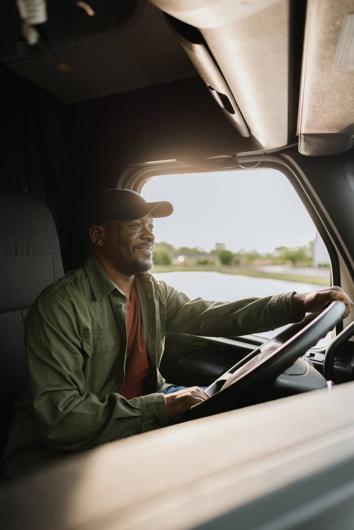 Person driving a truck, smiling. Wearing a casual shirt and cap. Image associated with Work & Money in a professional context