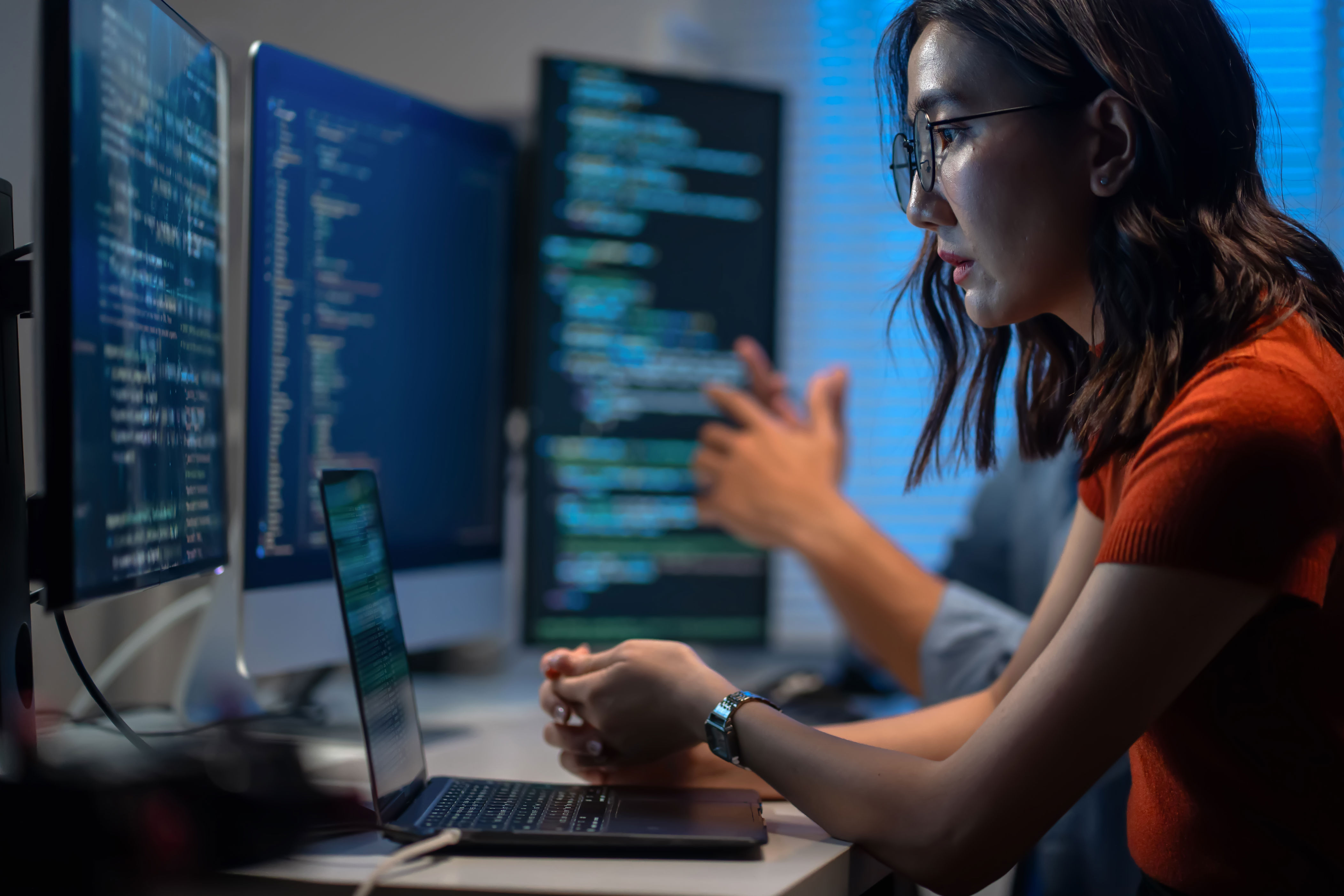 Woman focused on coding at a desk with multiple screens displaying code, collaborating with a colleague in an office setting