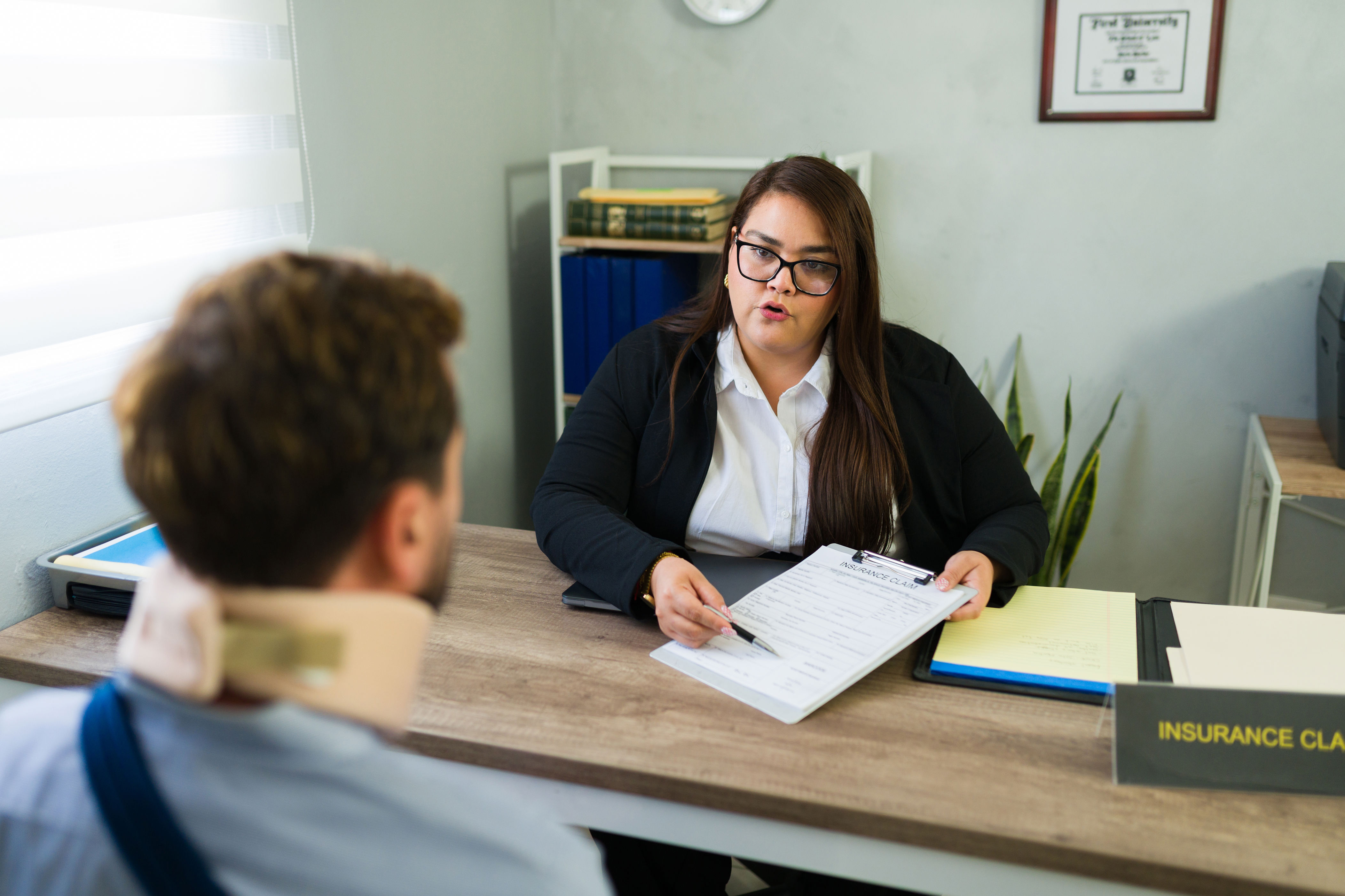 Person with neck brace consults with an insurance agent across a desk. The agent is holding a document and pen, ready to assist