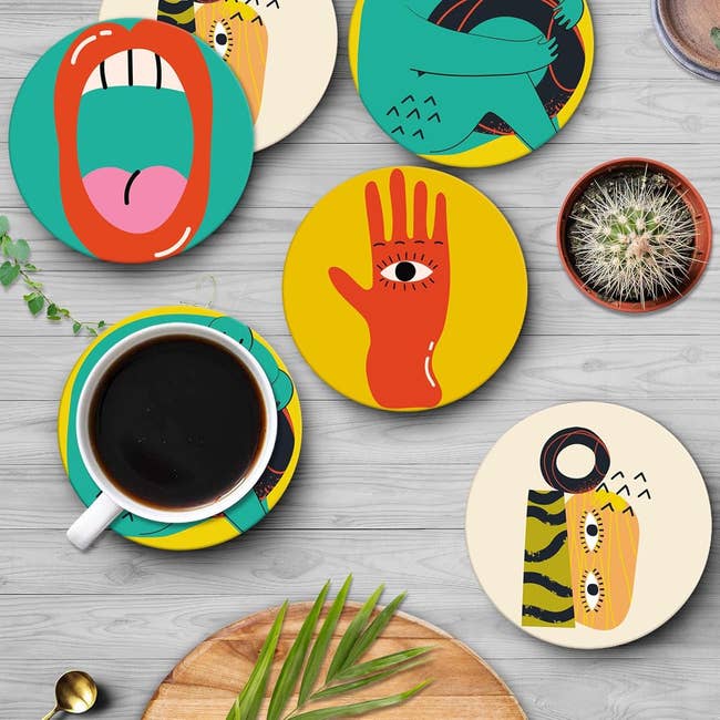 Artistic coasters with playful illustrations including a hand, mouth, and abstract designs, placed on a table with coffee and a cactus