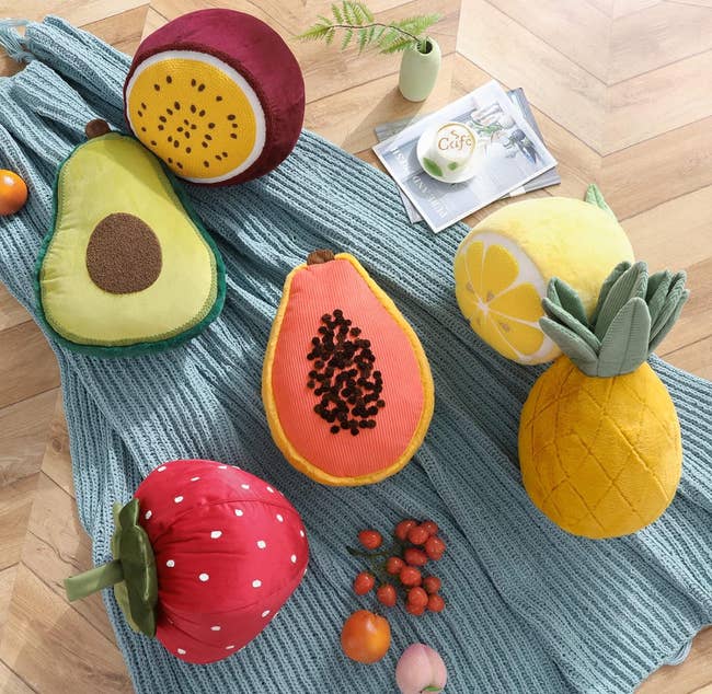 Plush fruit pillows, including avocado, papaya, pineapple, lemon, passion fruit, and strawberry, arranged on a blue knitted blanket