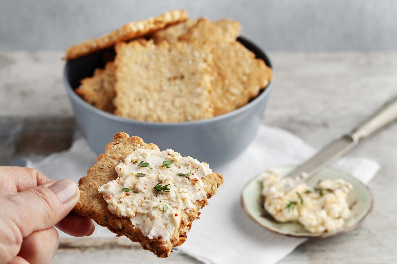 Hand holding a cracker with cheese spread; a bowl of crackers and more cheese in the background