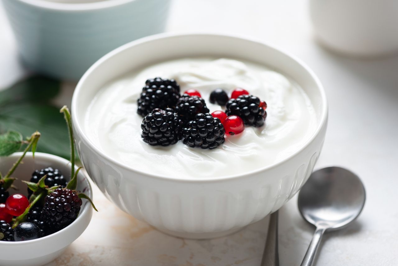A bowl of yogurt topped with blackberries and red berries alongside a spoon and another small bowl with blackberries