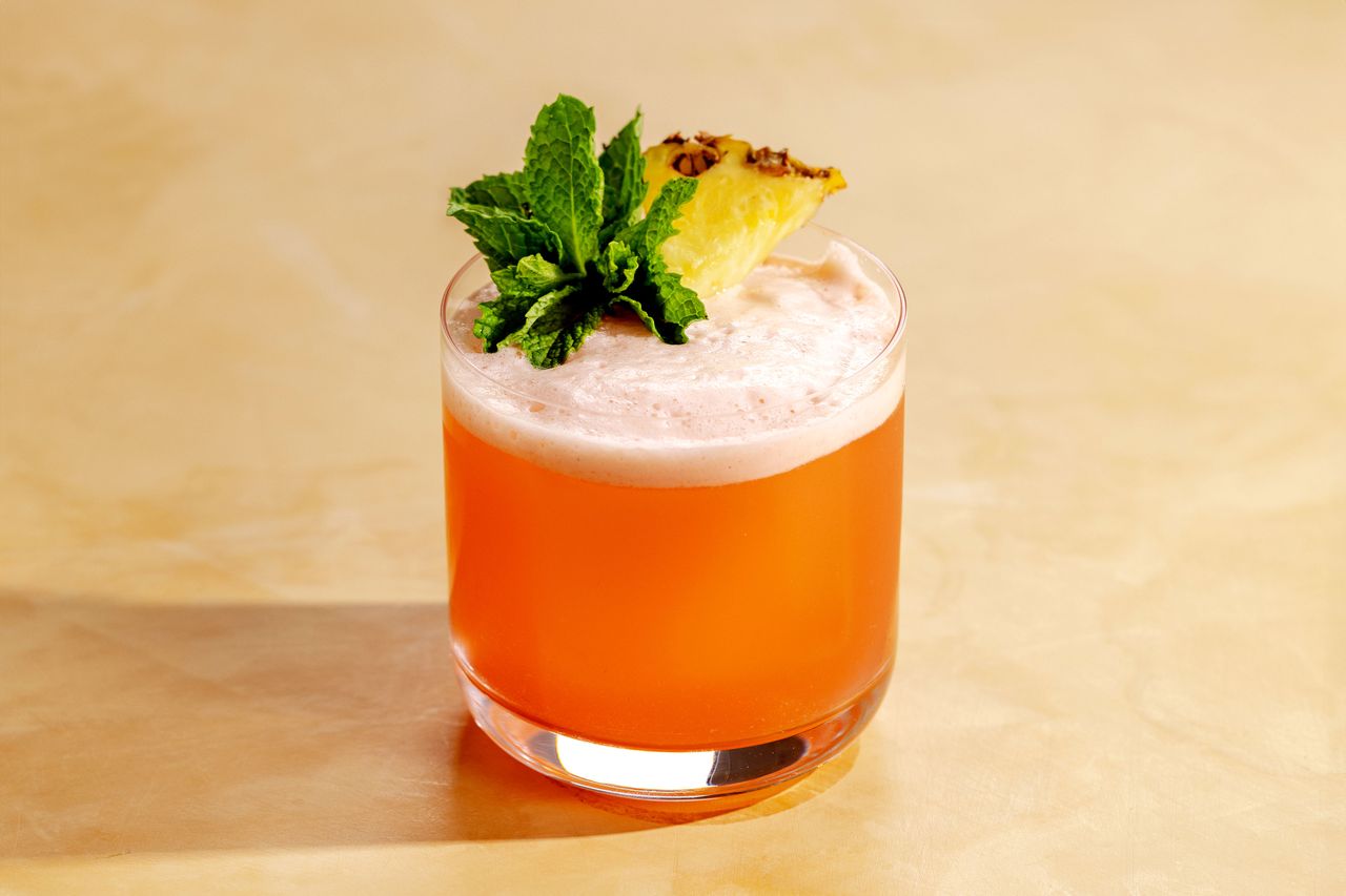 A jungle bird cocktail on a tan surface with a mint sprig and pineapple garnish.