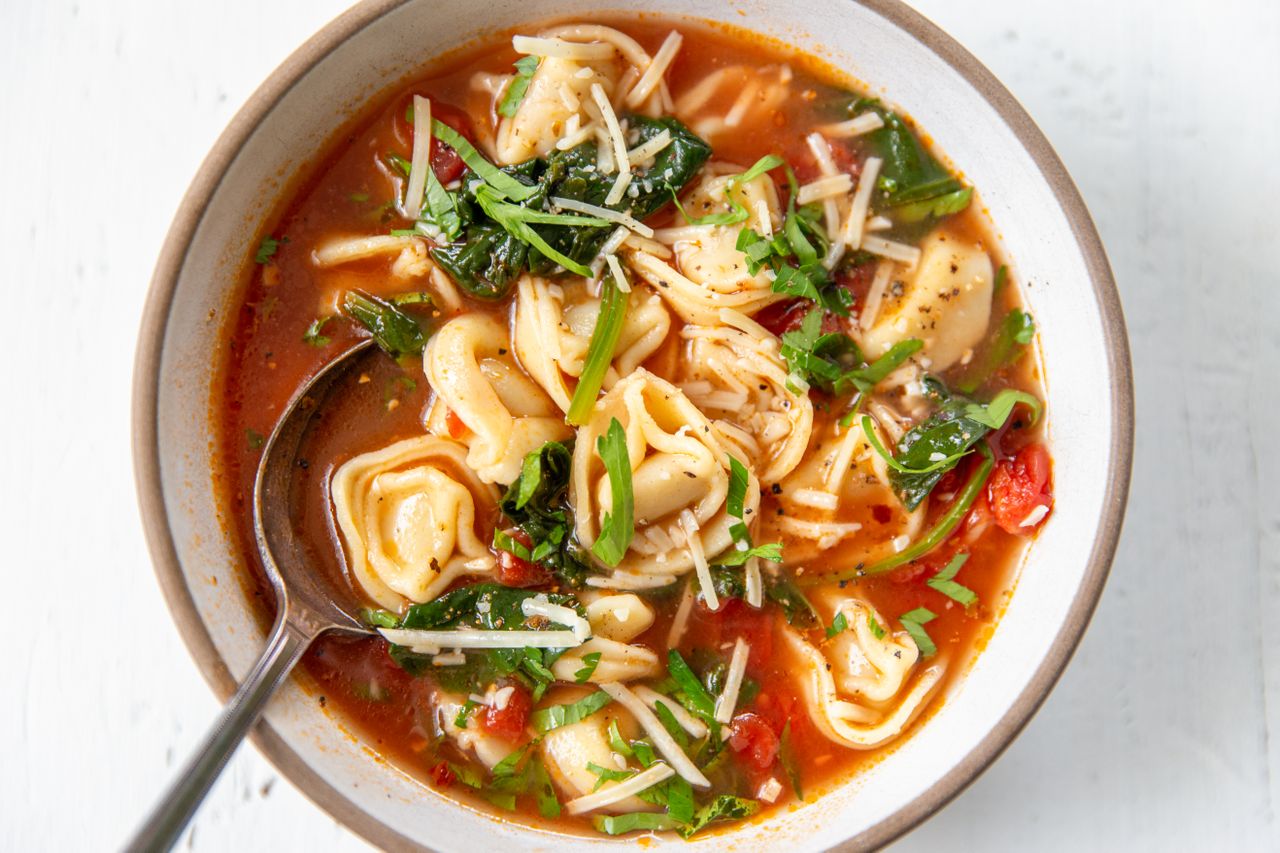 A serving of tortellini soup with a tomato based broth in a bowl, topped with parmesan cheese, with a spoon in the bowl