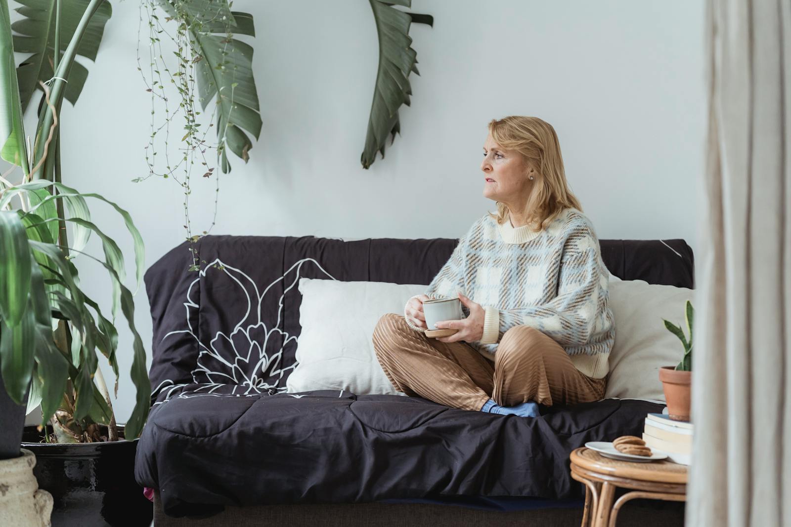 Full body of peaceful middle aged woman with blond hair in casual clothes drinking coffee and looking away thoughtfully while resting on sofa with crossed legs