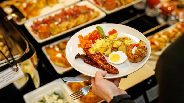 A hand holds a plate of breakfast food above dishes of food items at a breakfast buffet