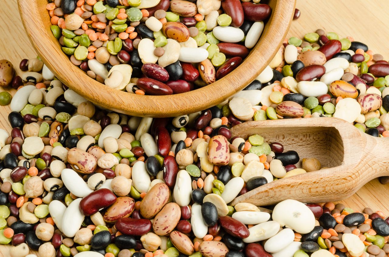 Various kinds of beans and lentils spilled on a surface with a wooden bowl and scoop.