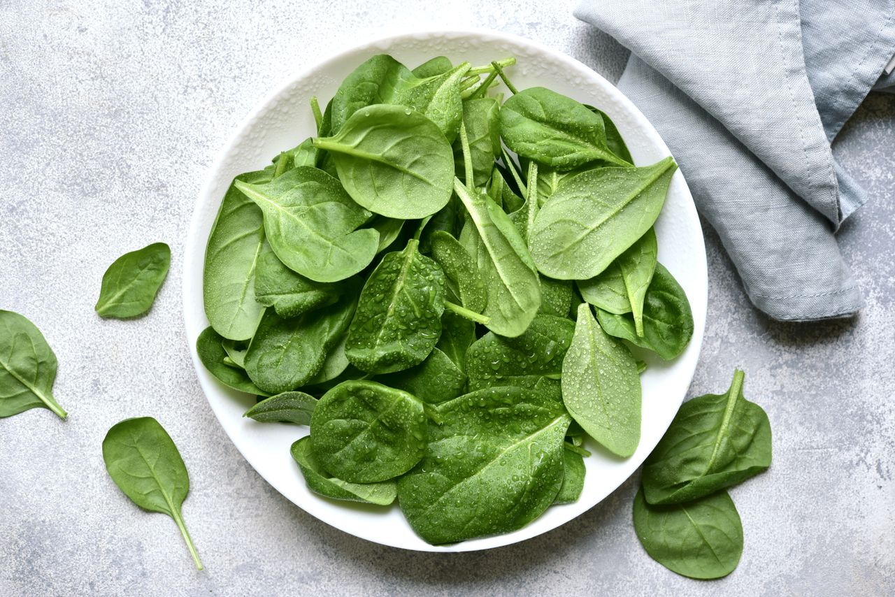 A plate of fresh spinach leaves on a gray surface with a cloth napkin in view.