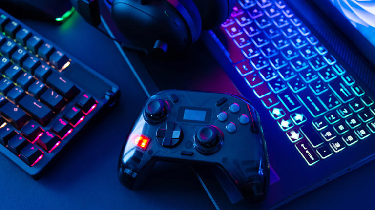 Gaming controller, headset, and keyboard next to laptop on a dark desktop