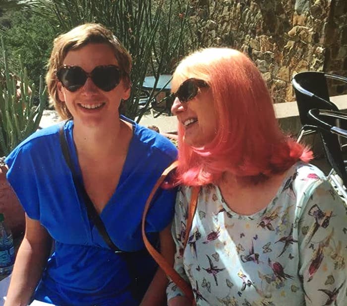 Two smiling people outdoors, wearing sunglasses; one in a blue top with a crossbody bag, the other in a floral blouse, with pink hair