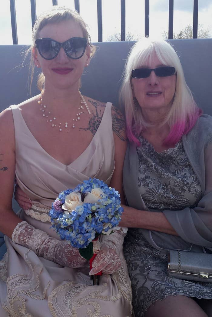 Two women sitting together. One in a satin gown with lace gloves holding a bouquet, the other in an embroidered dress. Both wearing sunglasses