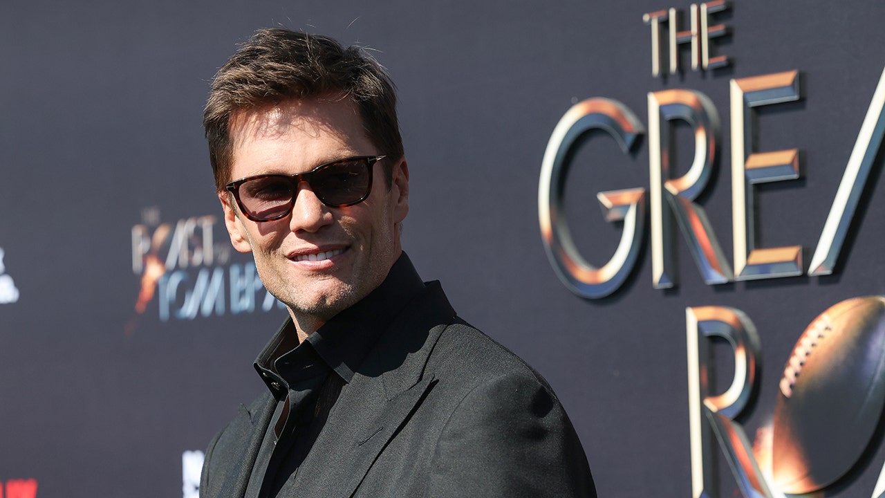 Tom Brady in a black suit and sunglasses looks dapper looking back at the camera at his roast