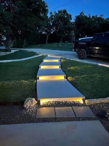 Pathway with illuminated steps at dusk, leading through a grassy area. A parked truck is visible to the right