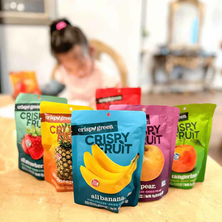 Brightly colored Crispy Green fruit snack pouches on a table with a blurred background of a young girl.